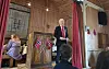 En mann taler fra scenen i et trepanelt rom med publikum ved bord og norske flagg.