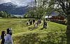 En gruppe mennesker står på en gresslette med fjell, fjord og trær rundt.