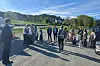 En gruppe festkledde mennesker står samlet utendørs på en parkeringsplass ved en grønn bakke i sollys.