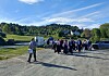 Mennesker samlet på en grusplass med biler, tilhenger og hus i et grønt, landlig område.