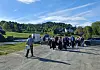 Mennesker samlet på en grusplass med biler, tilhenger og hus i et grønt, landlig område.