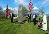 Flere personer står ved et steinmonument i en park med norske flagg og grønt gress.