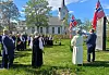 Personer står samlet på plenen foran en kirke med norske flagg og blå himmel i bakgrunnen.