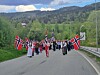 Barnetog med norske flagg på en landevei i grønt landskap med fjell bak.