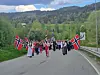 Barnetog med norske flagg på en landevei i grønt landskap med fjell bak.