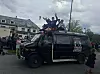 En svart varebil med folk stående på taket under en skyet himmel, omgitt av andre kjøretøy og tilskuere.