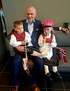 En mann sitter med to små barn i røde og svarte festdrakter på en benk innendørs.