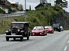 Flere biler, blant dem en mørk klassisk bil og en rød sportsbil, kjører langs en vei ved hus og trær.