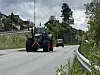Stor, 17. mai-pyntet traktor på vei med grønn natur, hus og en bil i bakgrunnen.