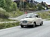Beige klassisk bil pyntet med norsk flagg, på en vei med hus i bakgrunnen.