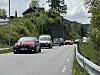 En bil pyntet med norske flagg kjører foran flere (stort sett?) pyntede biler på en svingete vei med skog og hus i bakgrunnen.
