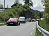 En bil pyntet med norske flagg kjører foran flere (stort sett?) pyntede biler på en svingete vei med skog og hus i bakgrunnen.