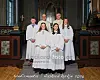 Fire konfirmanter i hvite kapper står foran alteret med to geistlige bak dem i en kirke.