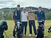 Fire golfspillere står på en golfbane med traller og bager foran åser og skog.