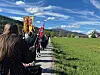 Mennesker går i 17. maitog langs en vei med faner ved et grønt jordbrukslandskap og fjell i bakgrunnen.