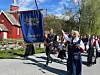Personer i bunad går i prosesjon forbi en rød kirke med en blå fane i front.
