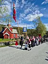 Prosesjon med norske flagg og bunadskledde deltakere foran en rød kirke på en solrik dag.