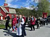 Korpsmedlemmer marsjerer i prosesjon langs en vei ved rød kirke og trær i vårvær.