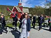 Et korps marsjerer langs veien med flagg, instrumenter og personer i mørke dresser.