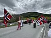 Barn og voksne i et 17. mai-tog med norske flagg på en vei foran fjell og skyer.