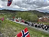 Folkemengde og bilkortesje med norske flagg langs en vei i et grønt landskap. Flere mennesker på biler.