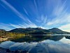 Fjell og blå himmel speiler seg i en stille innsjø med skyer over.