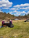 To personer spiser ved et piknikbord på en gresslette ved et vann med skog og blå himmel i bakgrunnen.
