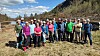 En gruppe mennesker står samlet på en grasbevokst elvebredd med fjell og skog i bakgrunnen.