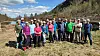 En gruppe mennesker står samlet på en grasbevokst elvebredd med fjell og skog i bakgrunnen.