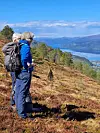 To turgåere med sekker står på en fjellskråning og ser utover fjord og dal under blå himmel.