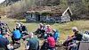 Flere personer sitter i en gruppe utenfor en liten steinhytte i et gresskledd landskap.