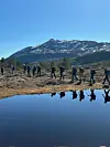 Mennesker går i rekke over et åpent område foran et snødekt fjell og et speilblankt vann.