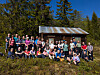 Stor gruppe personer poserer foran en liten hytte ved skogkanten.
