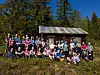 Stor gruppe personer poserer foran en liten hytte ved skogkanten.