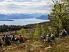 Personer sitter i gress og busker ved skogkanten med utsikt mot en innsjø og snødekte fjell.