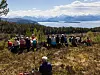Gruppe mennesker sitter på et utsiktspunkt med fjell, skog og vann i bakgrunnen.