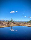 Speilblankt vann foran et åpent fjellandskap med blå himmel, små skyer og fjerne fjell.