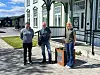 Tre personer står foran en hvitmalt bygning med grønne detaljer og en liten informasjonstavle ved inngangen.