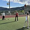 Tre personer på en bane spiller med en fargerik ball foran fjell og en bygning.