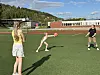 Tre barn spiller ball på en kunstgressbane med skolebygning og åser i bakgrunnen.