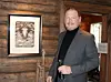 Mann i grå dress står i et rom med tømmervegger, et innrammet bilde og et glass i hånden.
