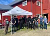 En gruppe personer står under et hvitt telt foran en rød låve på en gårdsplass.