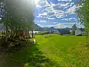 Grønn hage med plen, trær, hus og fjell under en lys himmel med skyer.
