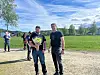 To menn går på en grusvei ved åpne jorder under blå himmel, med flere personer i bakgrunnen.