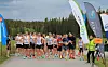 Gruppe løpere samlet ved startområdet med bannere og funksjonærer på en grusvei ved skog.