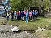 En gruppe personer står på en gressflate ved vannet, med en hvit svane foran dem.