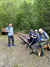 Tre personer sitter på tømmerstokker ved en skogssti, mens en fjerde står ved siden av med ryggsekk.