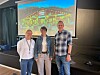 Tre personer står foran en presentasjonsskjerm med et bilde av en bygning og rødt logo-skilt.