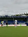 Fotballspillere på gressbanen foran en tribune med reklameskilt og overskyet himmel.