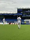 To fotballspillere på en grønn bane foran tomme tribuner og reklameskilt på et stadion.